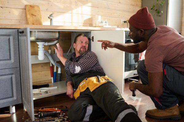 Mature plumber looking at young colleague pointing at pipe under metallic sink in kitchen cabinet while explaining the problem